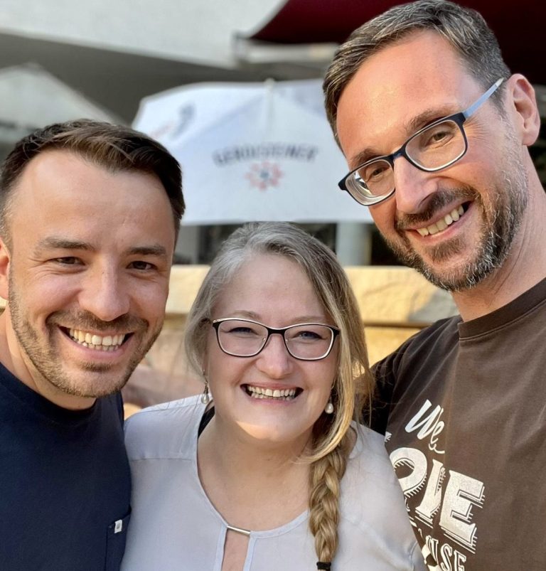 Drei Personen lächeln in einem Cafe, zwei Männer und eine Frau mit Brille.