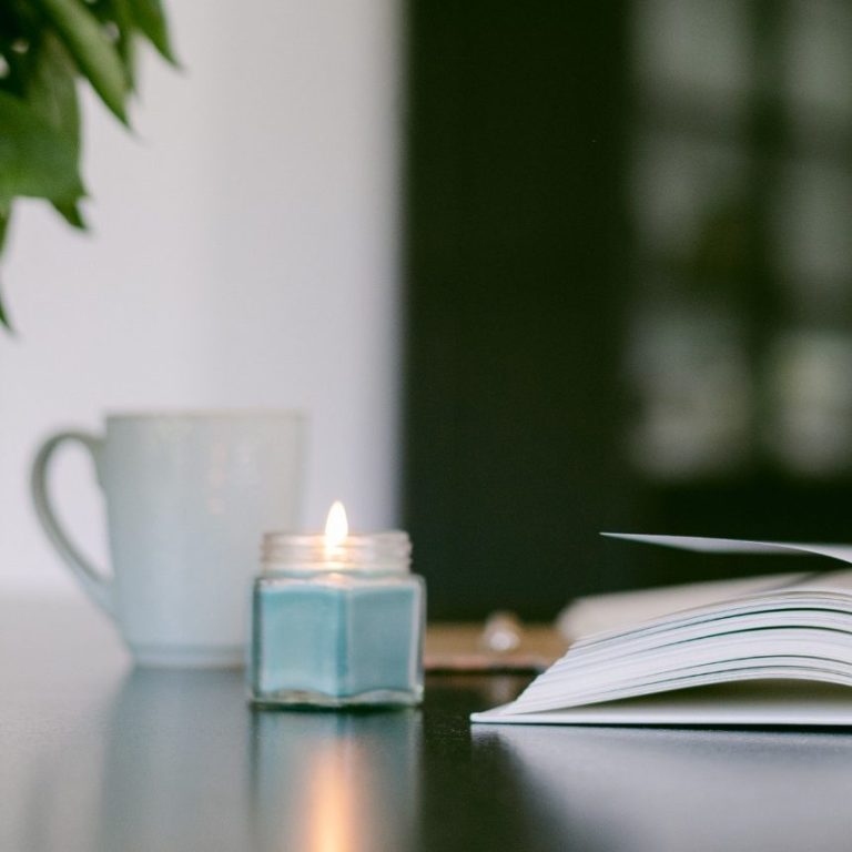 Tasse, brennende Kerze und offenes Buch auf einem Tisch, umgeben von Pflanzen.